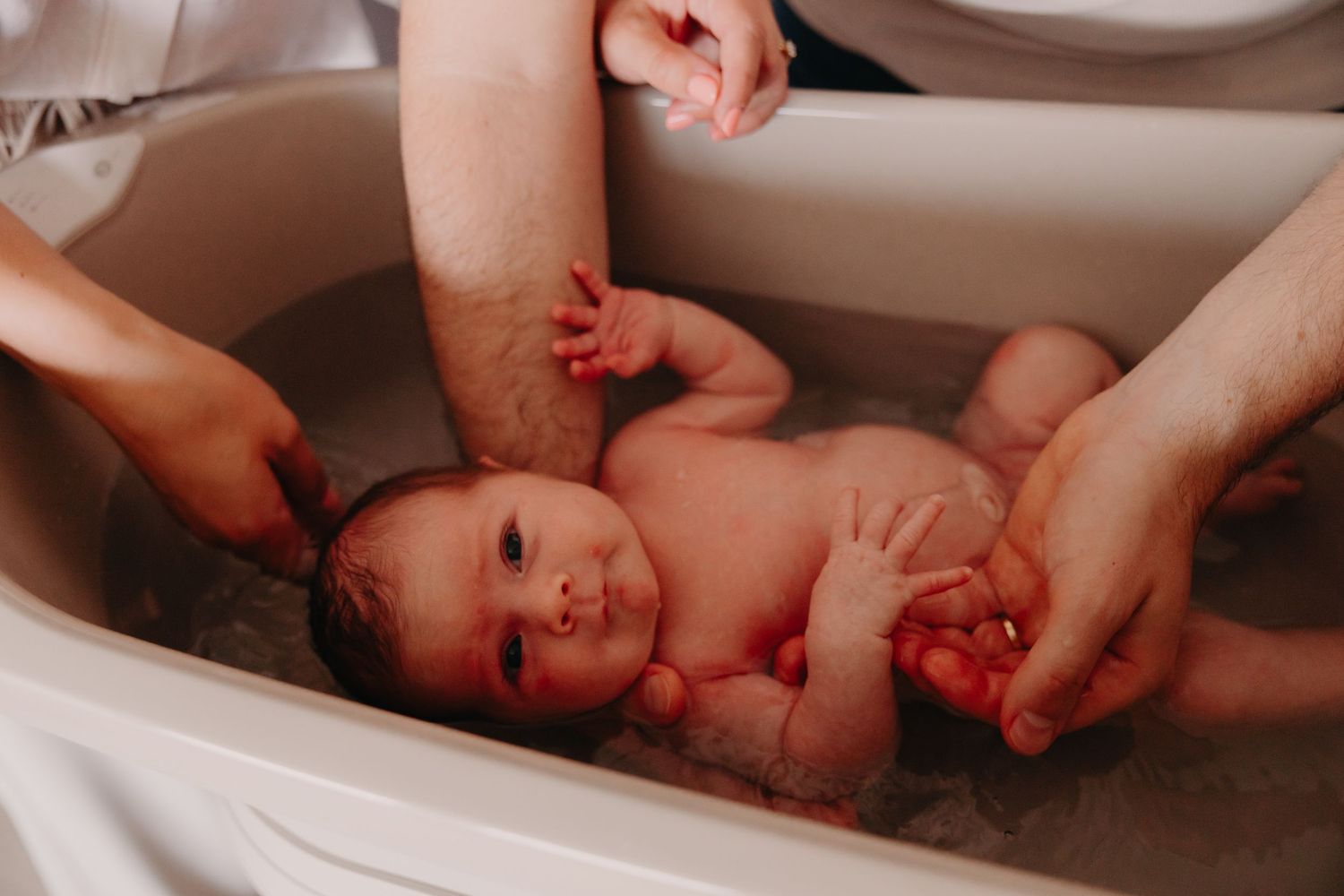 Newborn baby being gently bathed in a small white tub by parents.