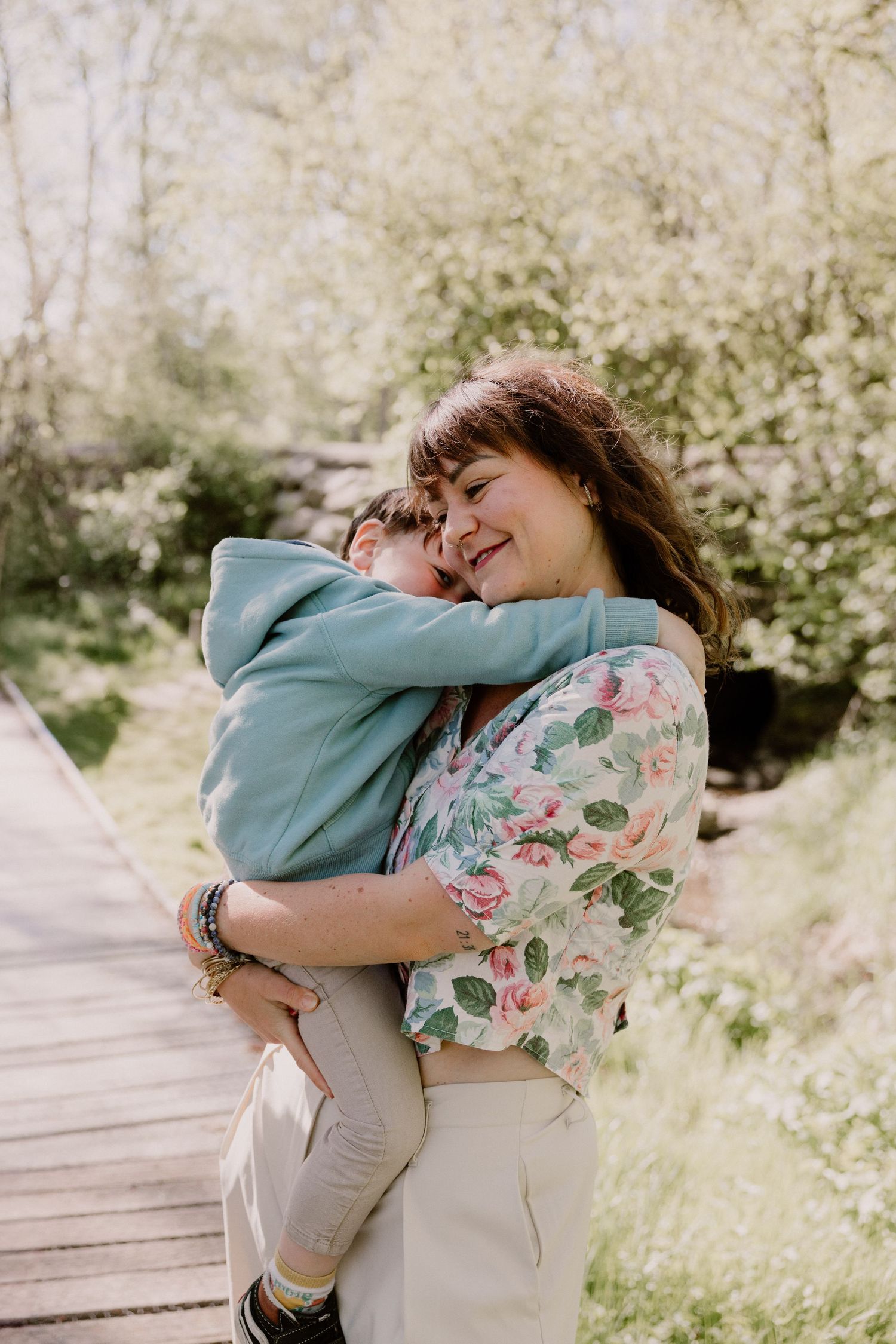Joyful mother in floral top hugging and lifting her young child outdoors in a sunny park.