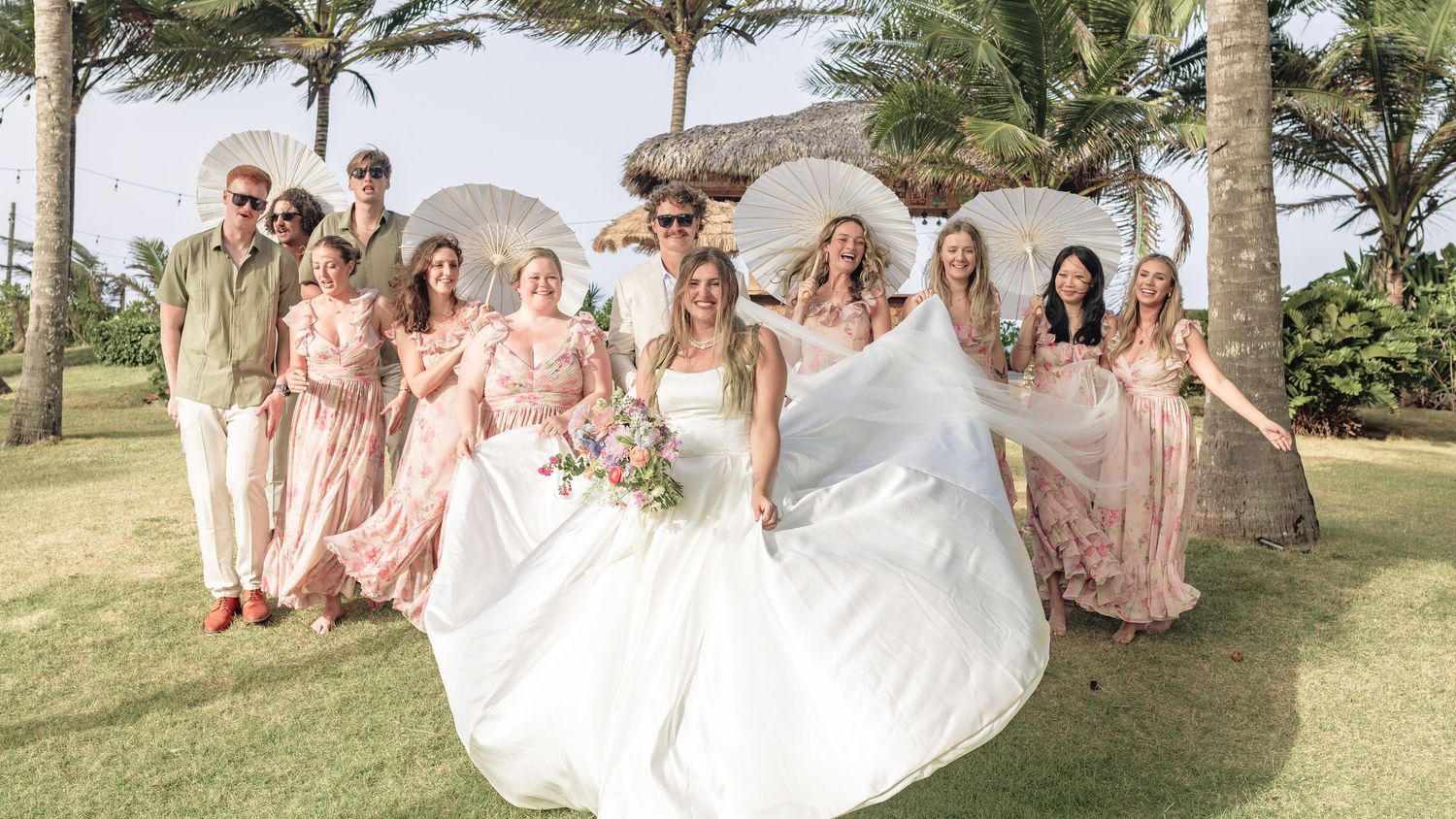 Bride in white gown poses with wedding party in blush pink dresses on tropical beach with palm trees.