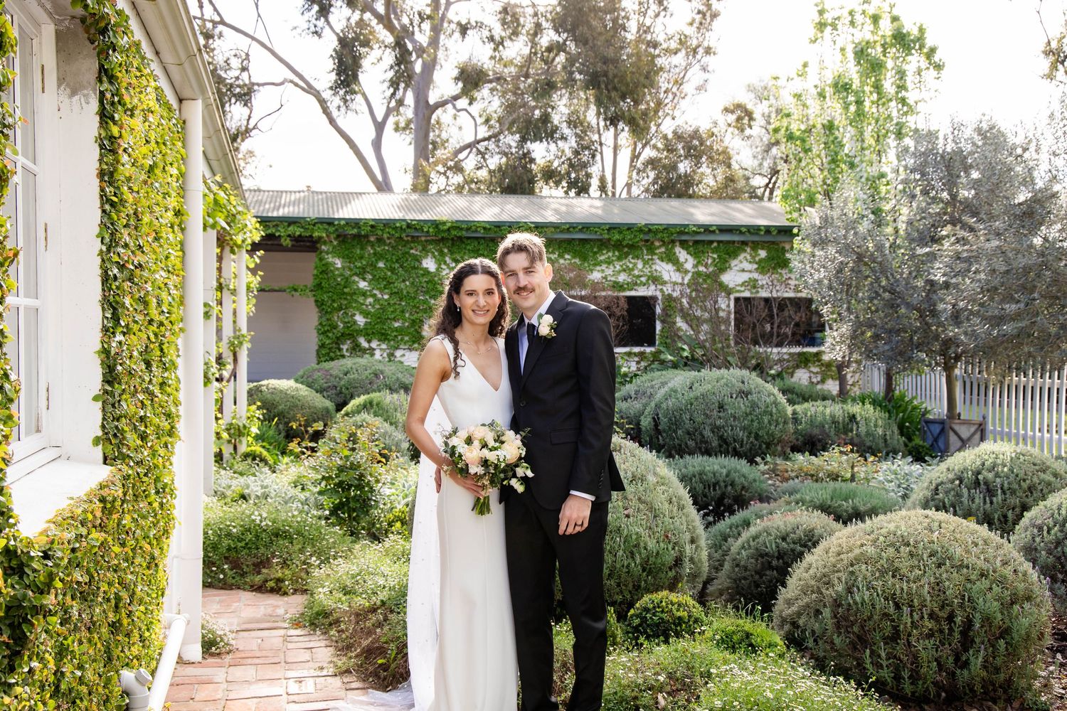 Bride and groom pose together in a lush garden with ivy-covered walls and manicured hedges on their wedding day.