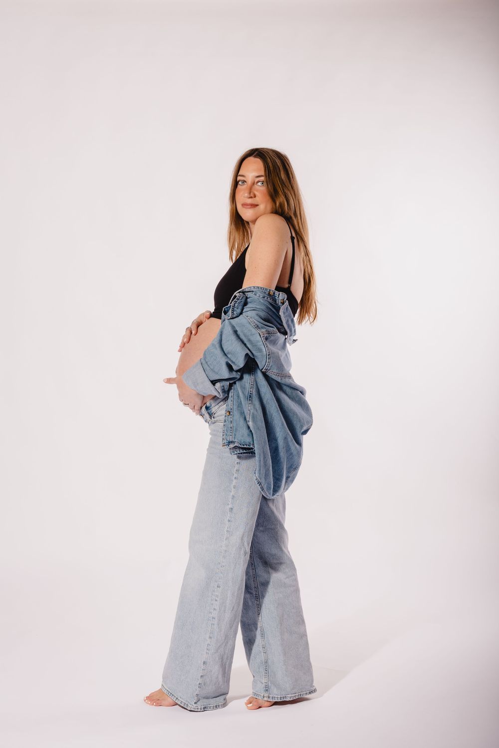 Pregnant woman in denim jacket and wide-leg jeans cradling her baby bump against a white background.