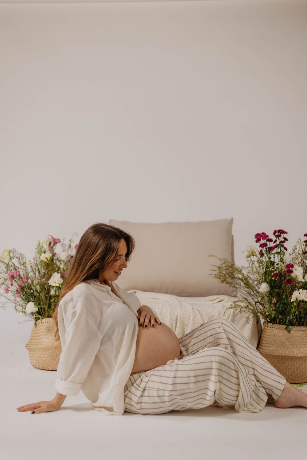 Pregnant woman sitting on floor leaning against chair with floral arrangements, wearing white shirt and striped pants.
