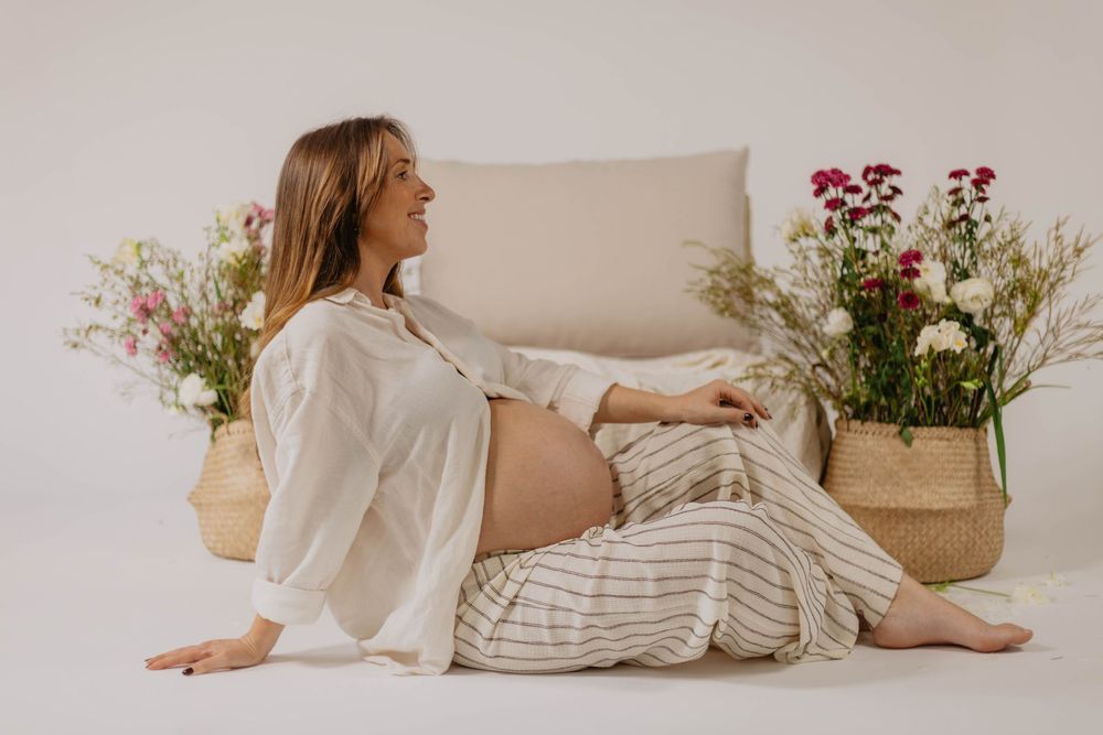 Pregnant woman in white blouse and striped pants sitting on floor surrounded by flowers in woven baskets.