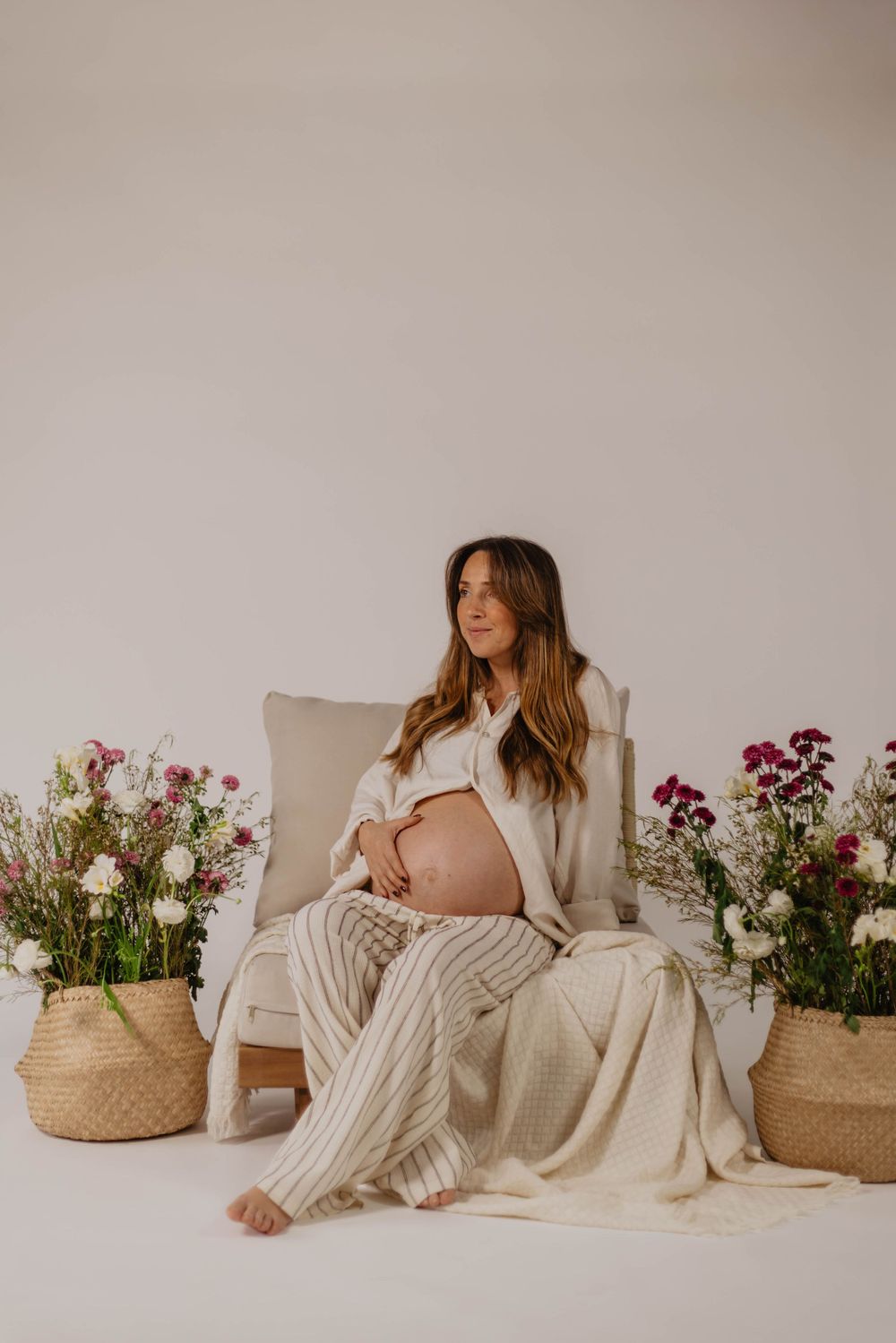 Pregnant woman sitting in chair surrounded by flowers in wicker baskets, wearing white shirt and striped linen pants.