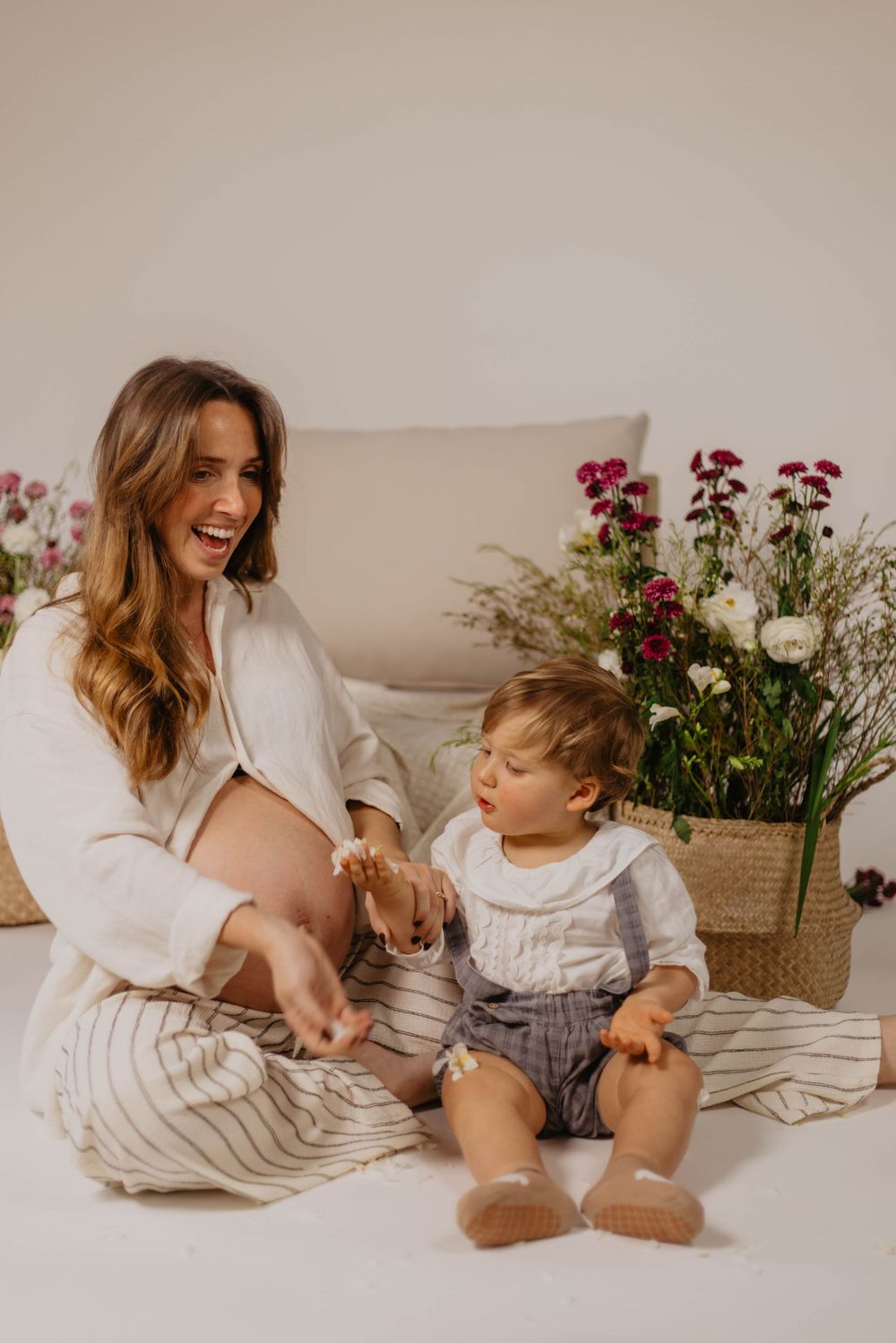 Pregnant woman laughing with toddler boy sitting on blanket surrounded by flowers in a bright studio setting.
