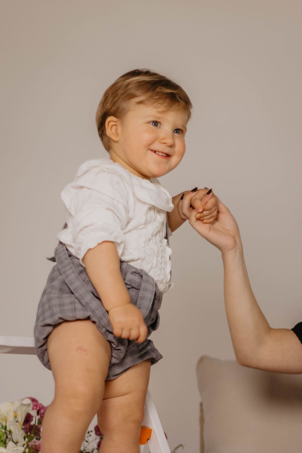 Happy toddler in white shirt and gray shorts smiling while holding an adult's hand indoors.