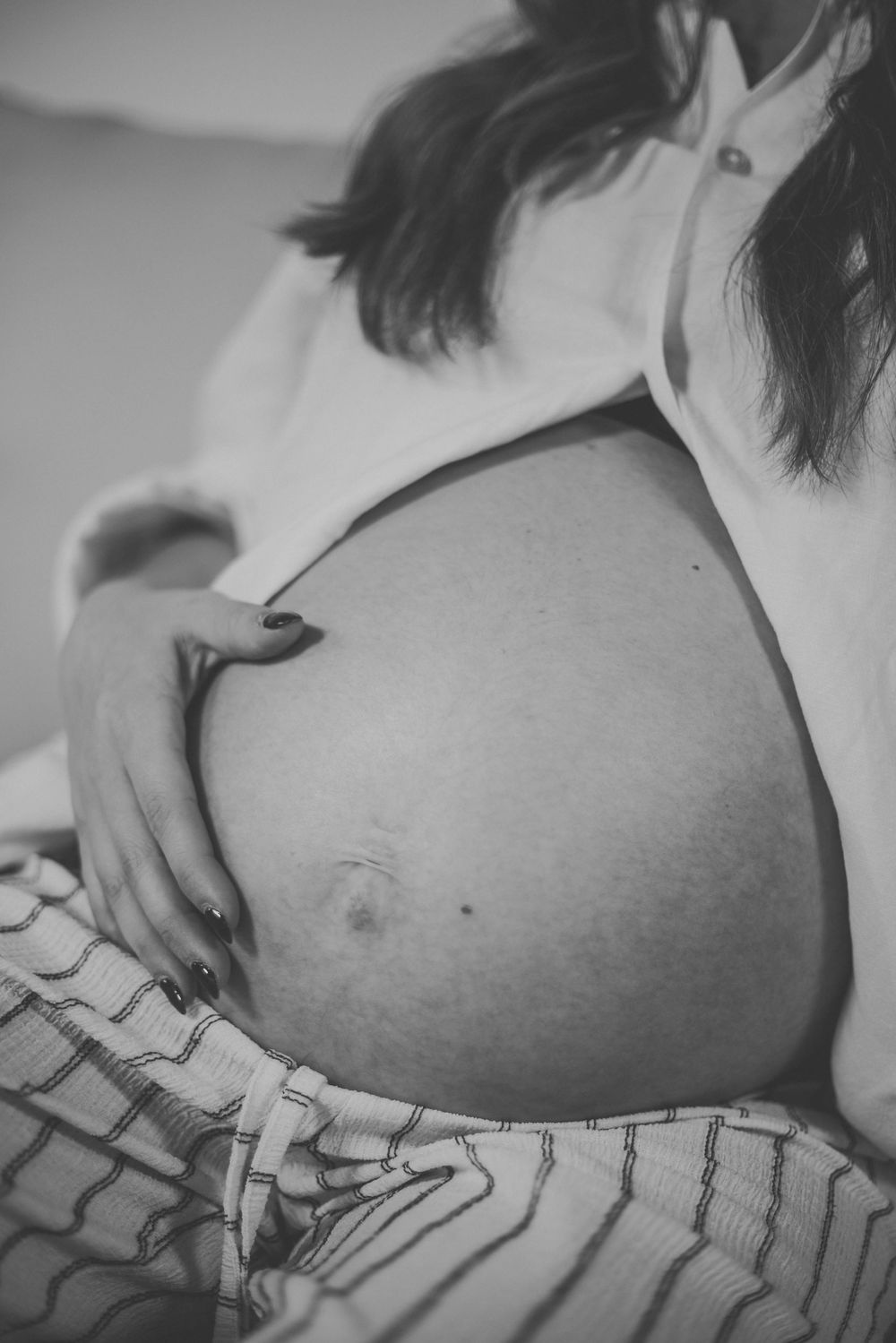 Black and white close-up of pregnant woman cradling baby bump, wearing open white shirt and striped pants.