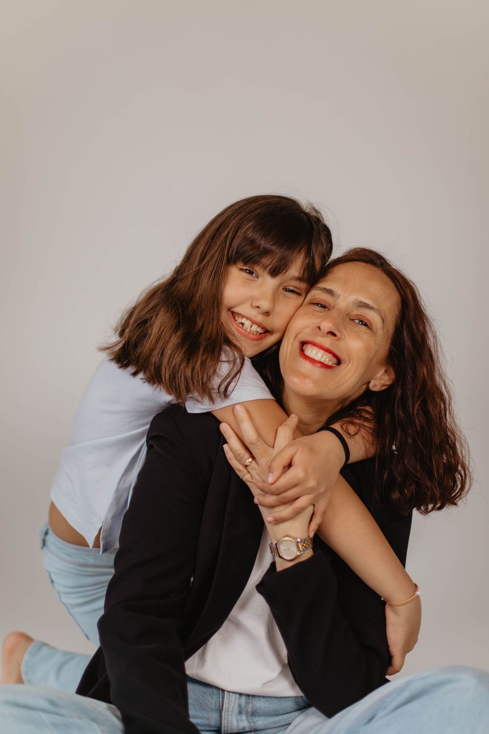 A joyful mother and daughter hugging and laughing together, both wearing black jackets and jeans on white background.