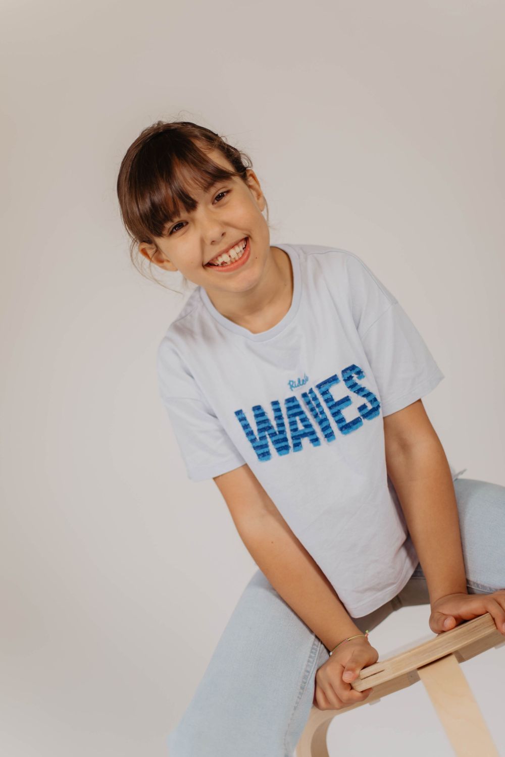 Smiling girl in white Waves t-shirt and jeans, sitting on wooden stool against light gray background.