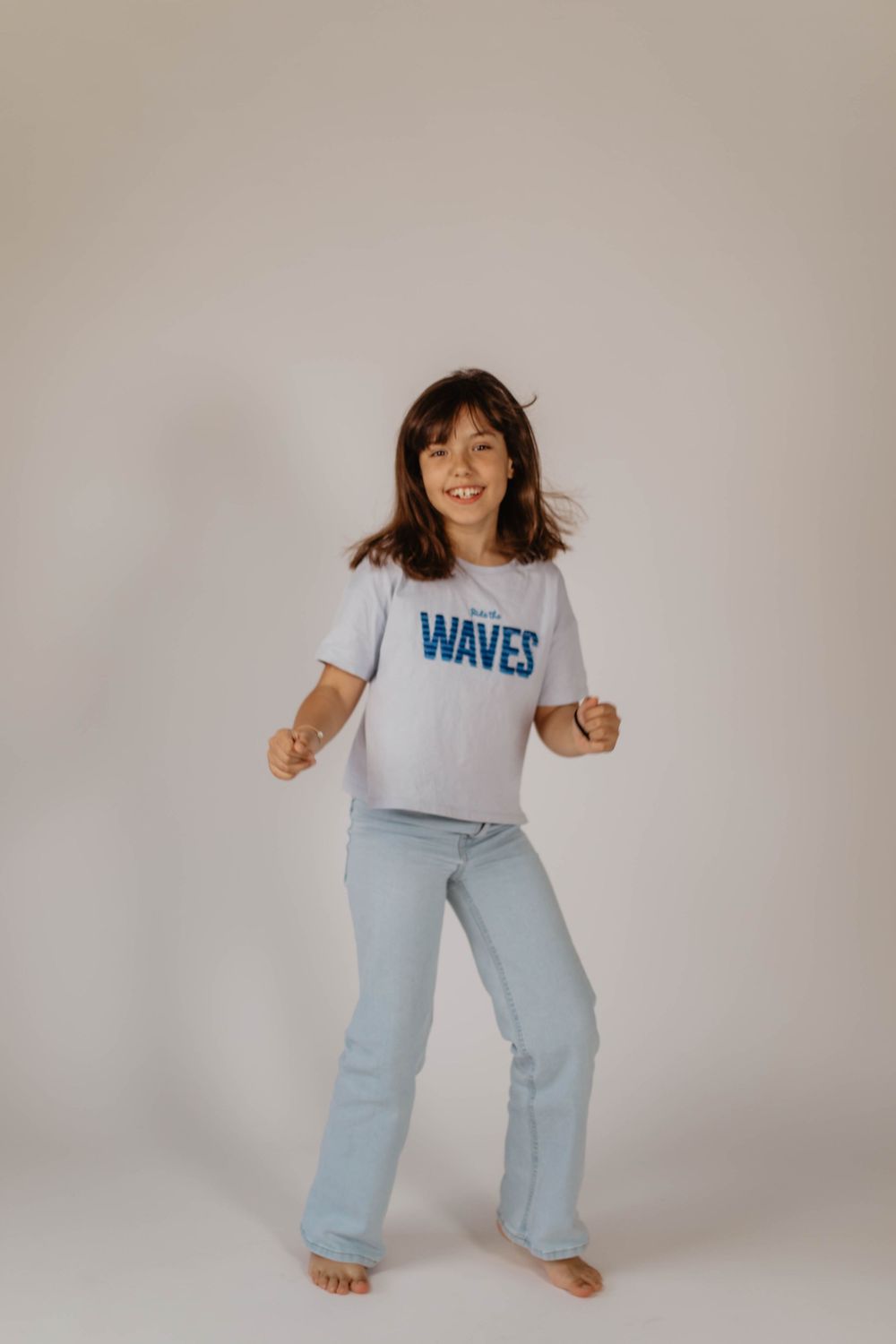 Young girl dancing in a white Waves graphic tee and light blue flare jeans, barefoot on a white background.