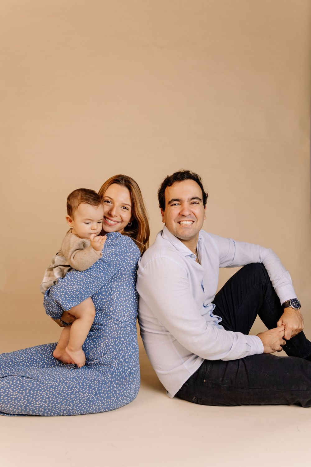 Smiling couple sitting back-to-back on floor with their baby in a warm beige studio family portrait.