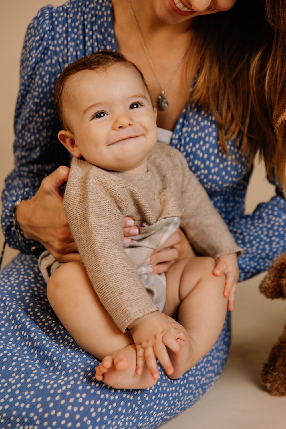 Adorable baby in a knit sweater smiling while being held by mother in a blue floral dress.
