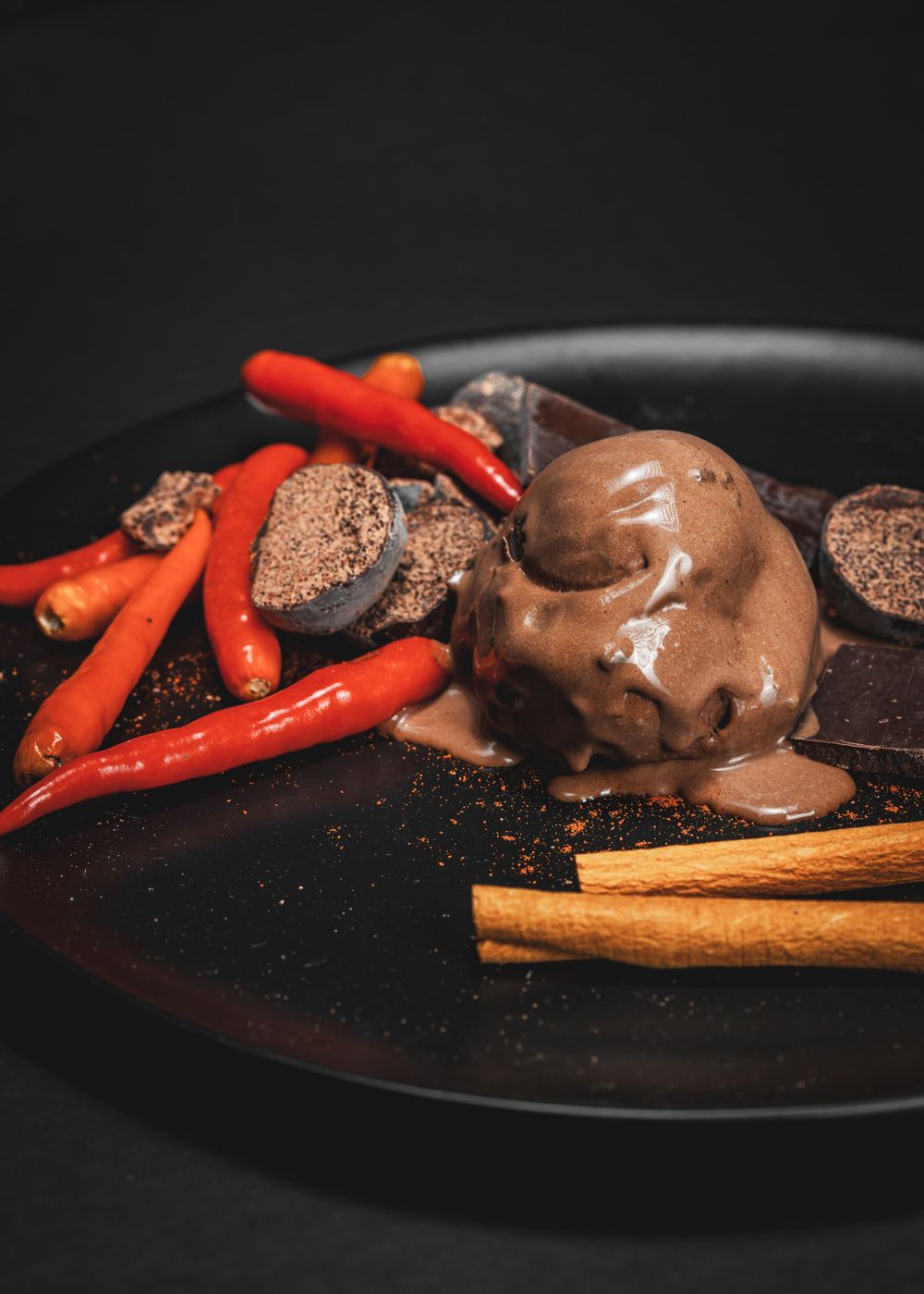 Dark chocolate ice cream scoop with chili peppers, cinnamon sticks, and cocoa powder on a black plate.