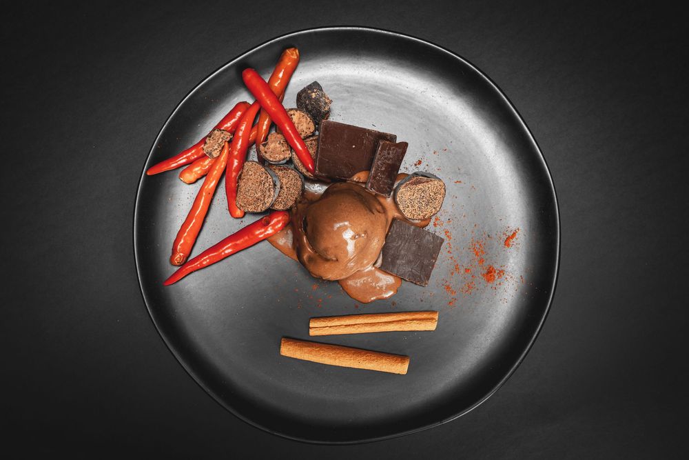 Chocolate ice cream scoop with cinnamon sticks, dark chocolate pieces, and red chili peppers on a dark plate.