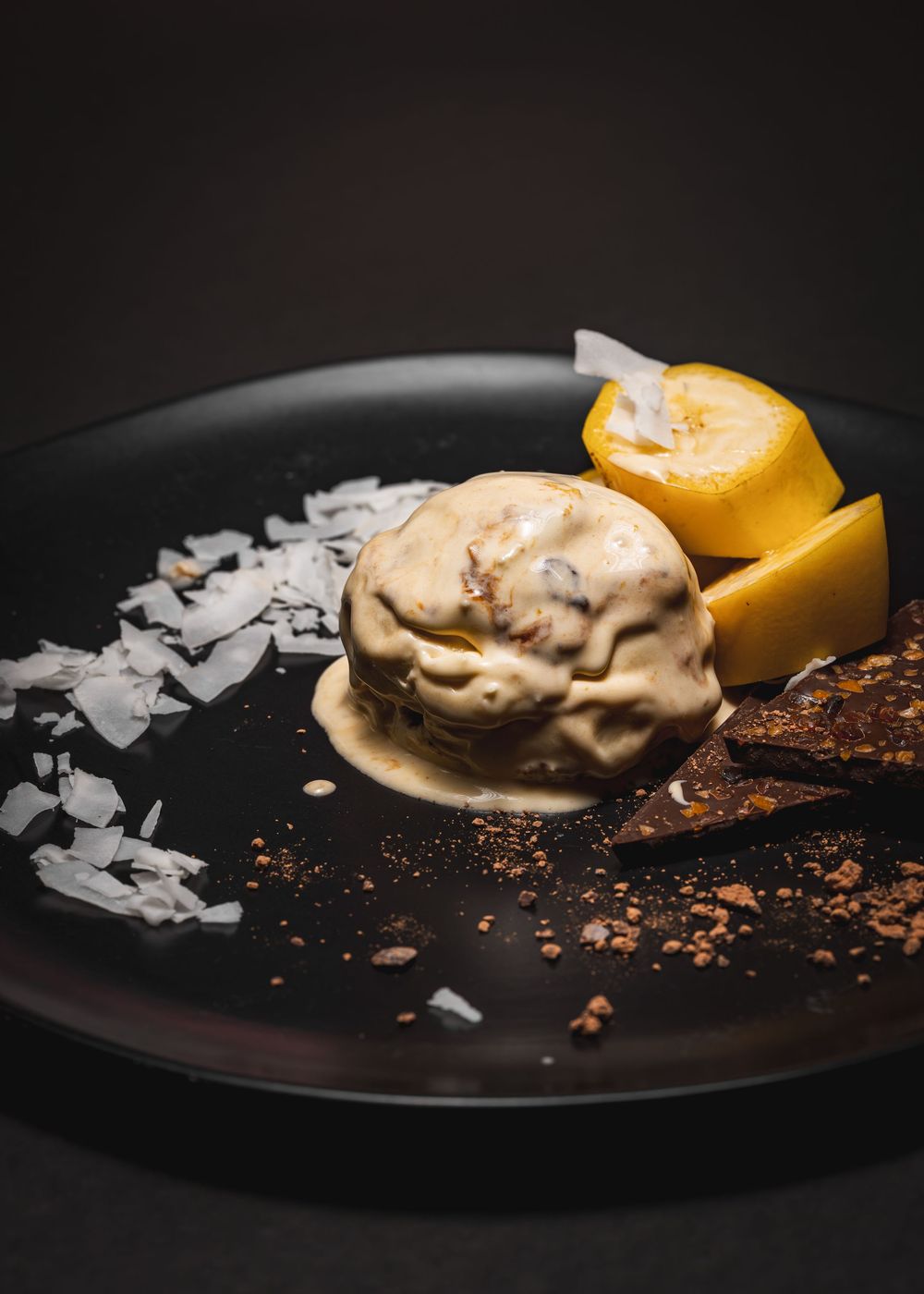 A scoop of caramel ice cream on a black plate with mango slices, coconut flakes, and dark chocolate shards.