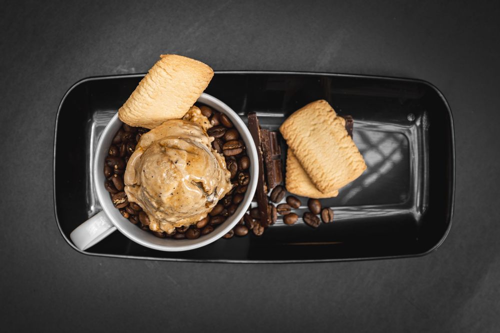 Coffee ice cream scoop in a mug with coffee beans and shortbread cookies on a black tray.