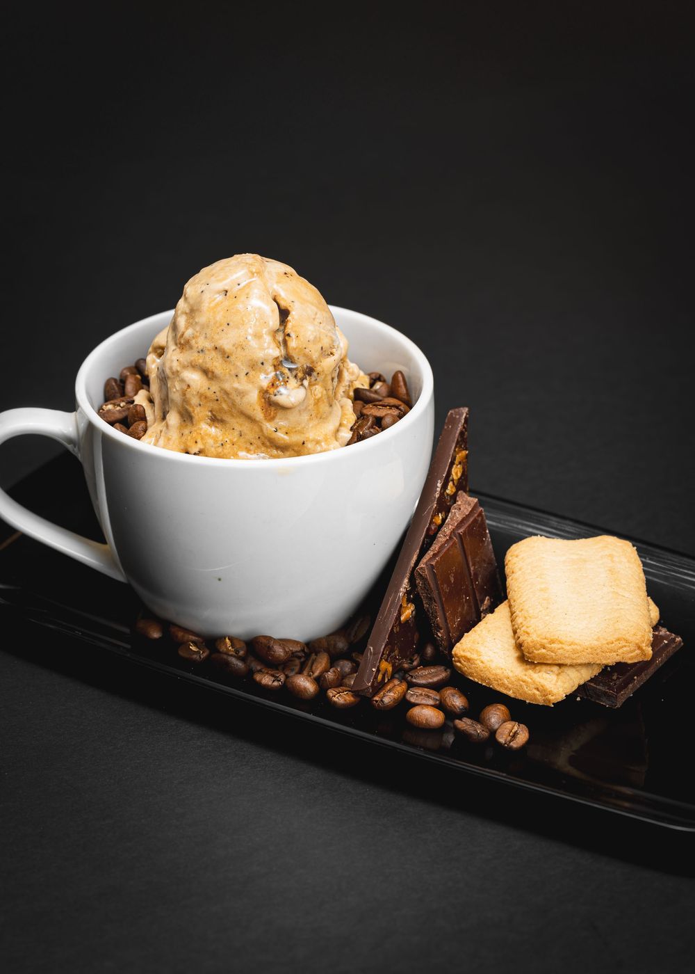 Coffee ice cream scoop in white mug with coffee beans, chocolate, and shortbread cookies on black tray.