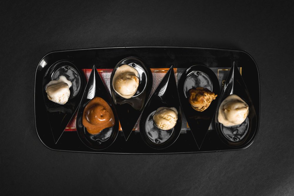 Elegant black serving tray with assorted ice cream scoops and sauces in individual spoon-shaped dishes on dark background.