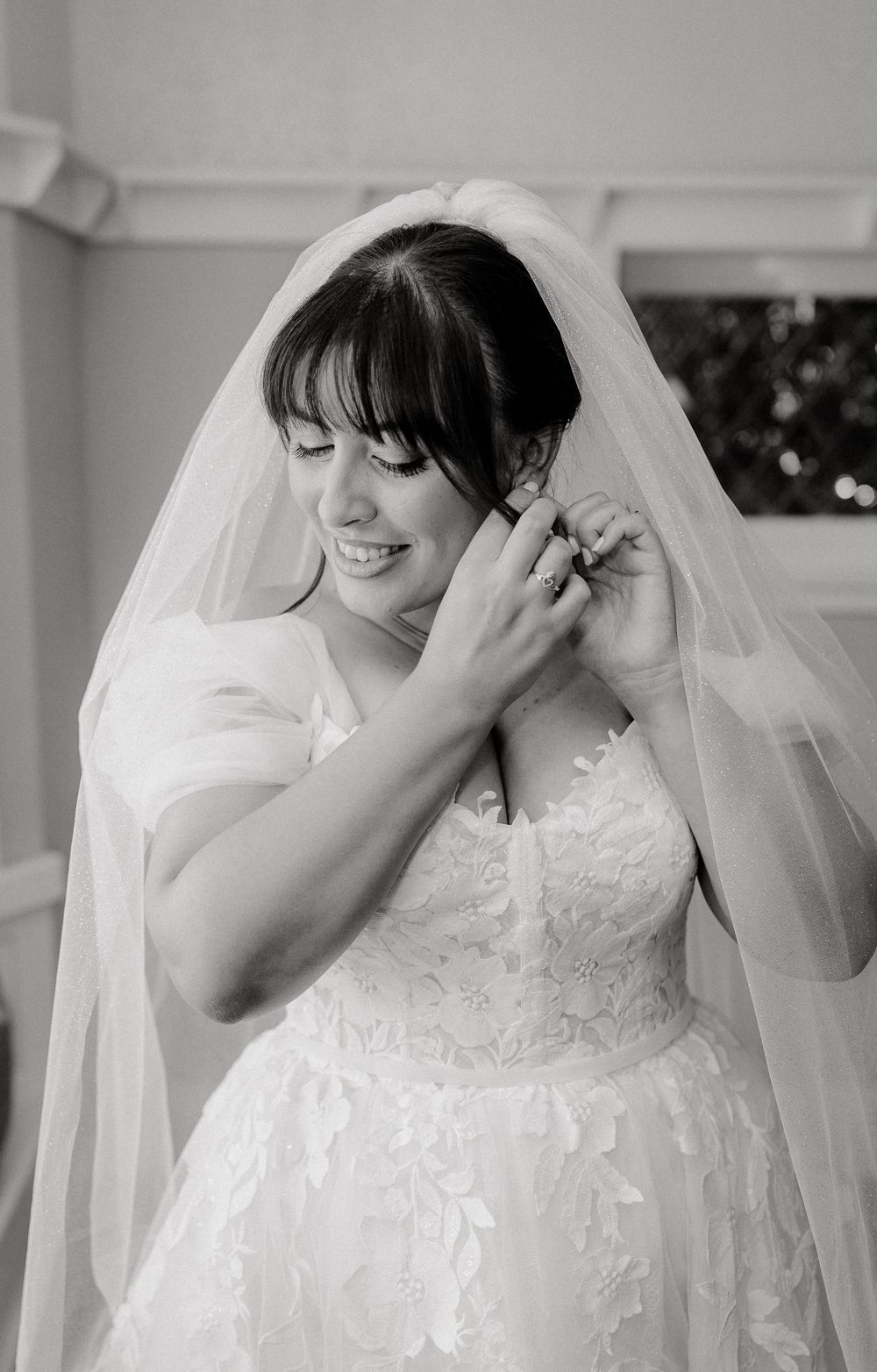 Smiling bride in lace wedding dress and veil adjusting her earring while getting ready on her wedding day.