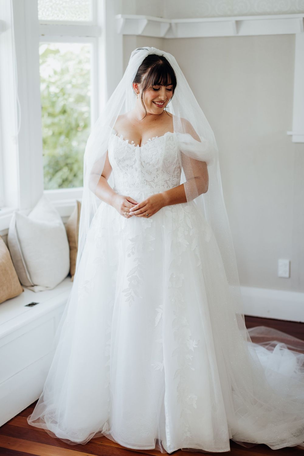 Smiling bride in an off-shoulder lace A-line wedding dress with a long veil standing by a window.