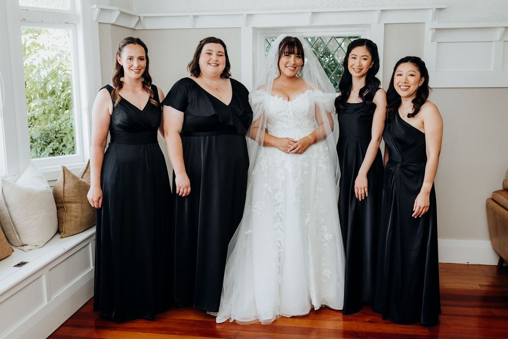 Bride in white lace gown poses with four bridesmaids in black dresses indoors.