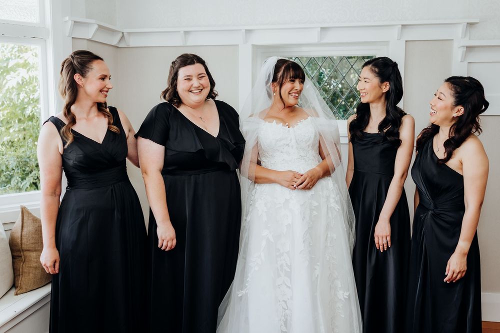Bride in white lace gown smiling with four bridesmaids in black dresses before wedding ceremony.