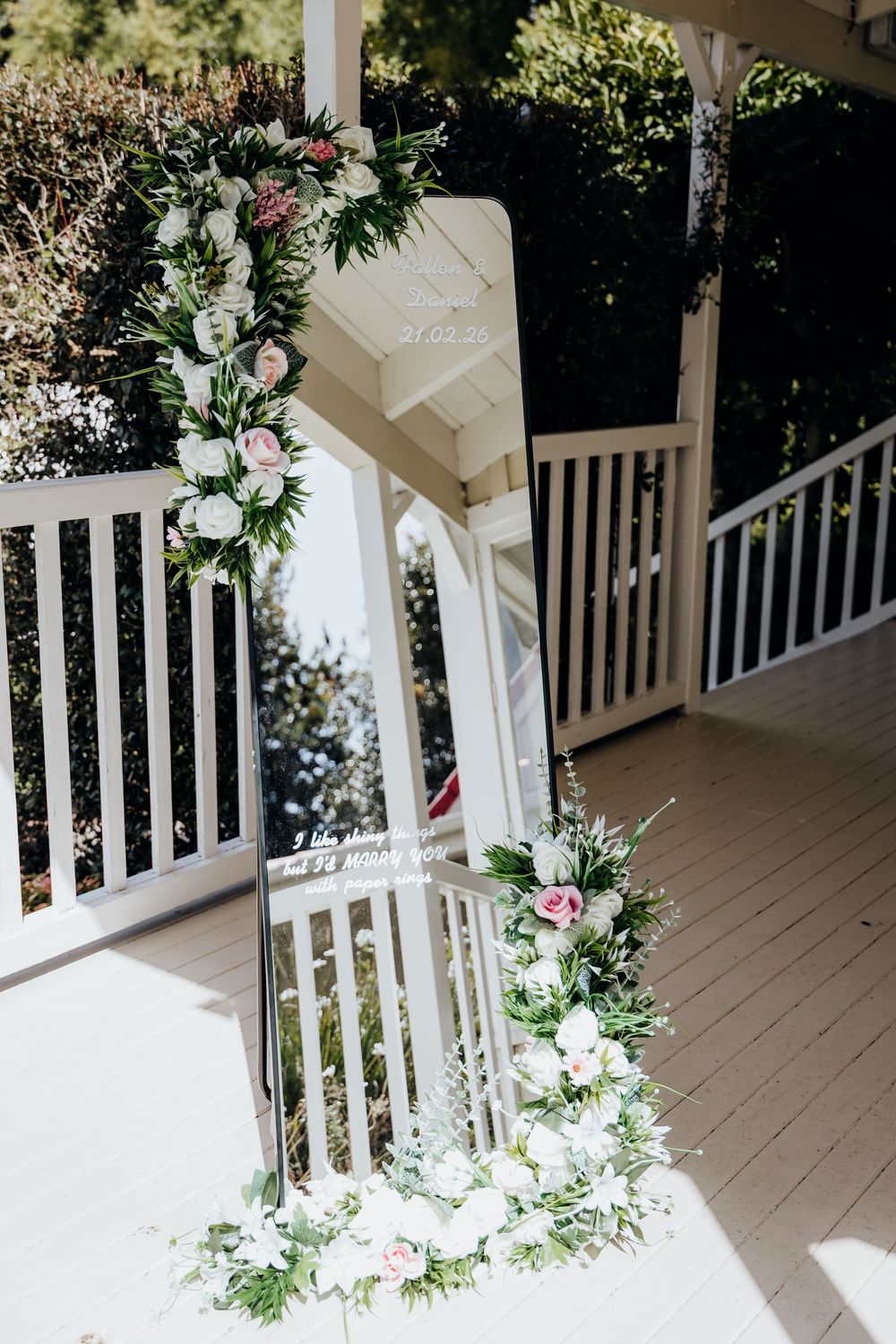 Elegant wedding arch with pink and white floral arrangements on a white porch, featuring a decorative mirror with script writing.