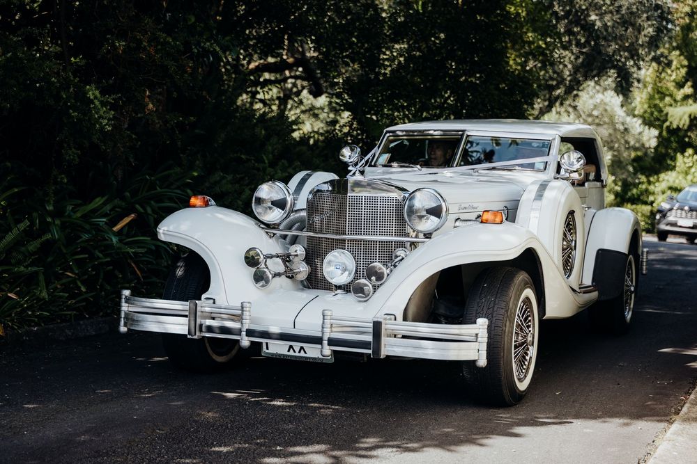 Classic white vintage Excalibur wedding car decorated with ribbons parked on a tree-lined driveway.