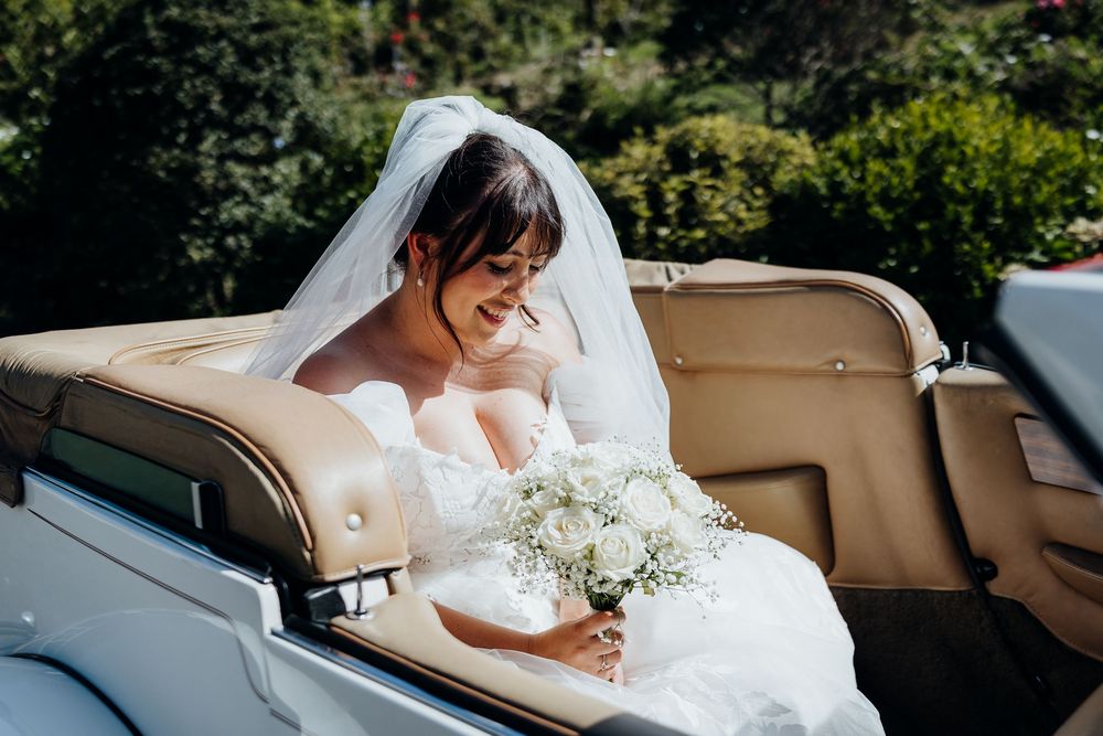 Beautiful bride in white gown and veil holding bouquet while seated in vintage wedding car.