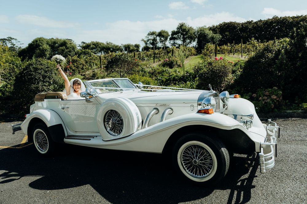 Bride waving from a classic white vintage Beauford convertible wedding car on a sunny day.
