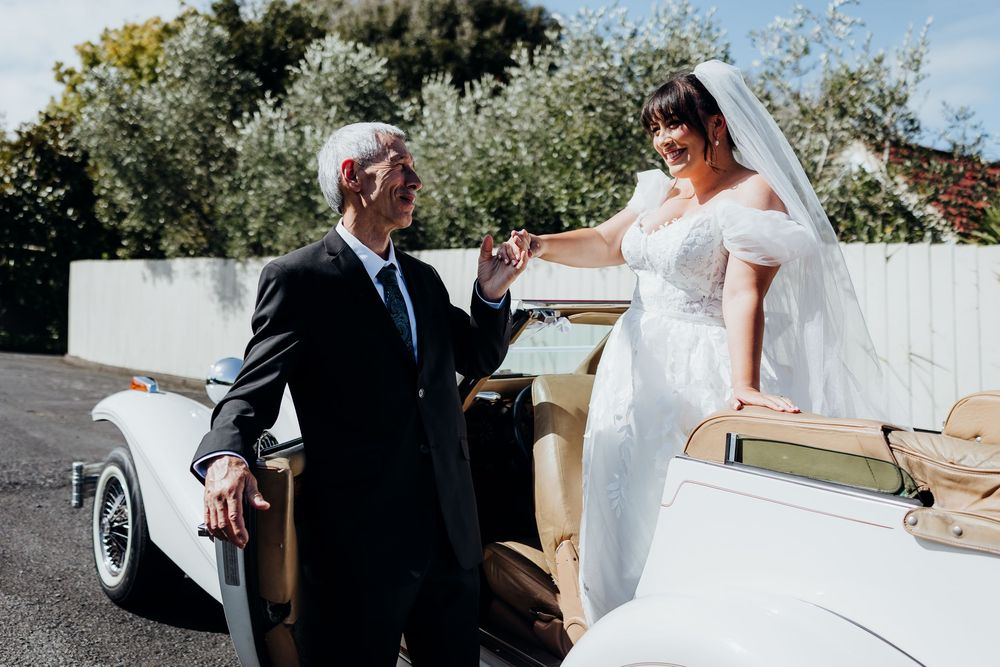 Bride in white dress and veil greeting older gentleman beside a classic vintage white wedding car.