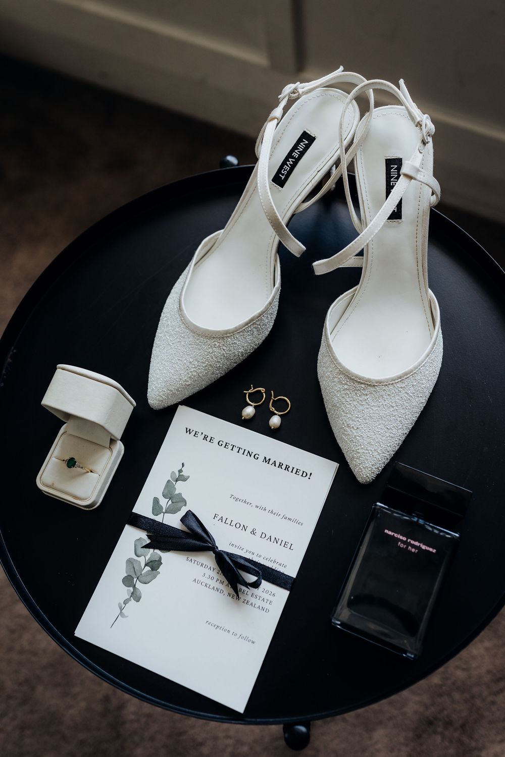Elegant bridal flat shoes, pearl earrings, engagement ring, and wedding invitation on black table.