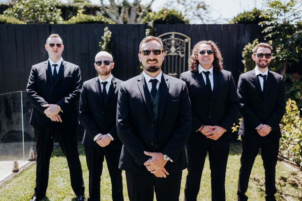 Five groomsmen in matching black suits and sunglasses posing outdoors before a wedding ceremony.