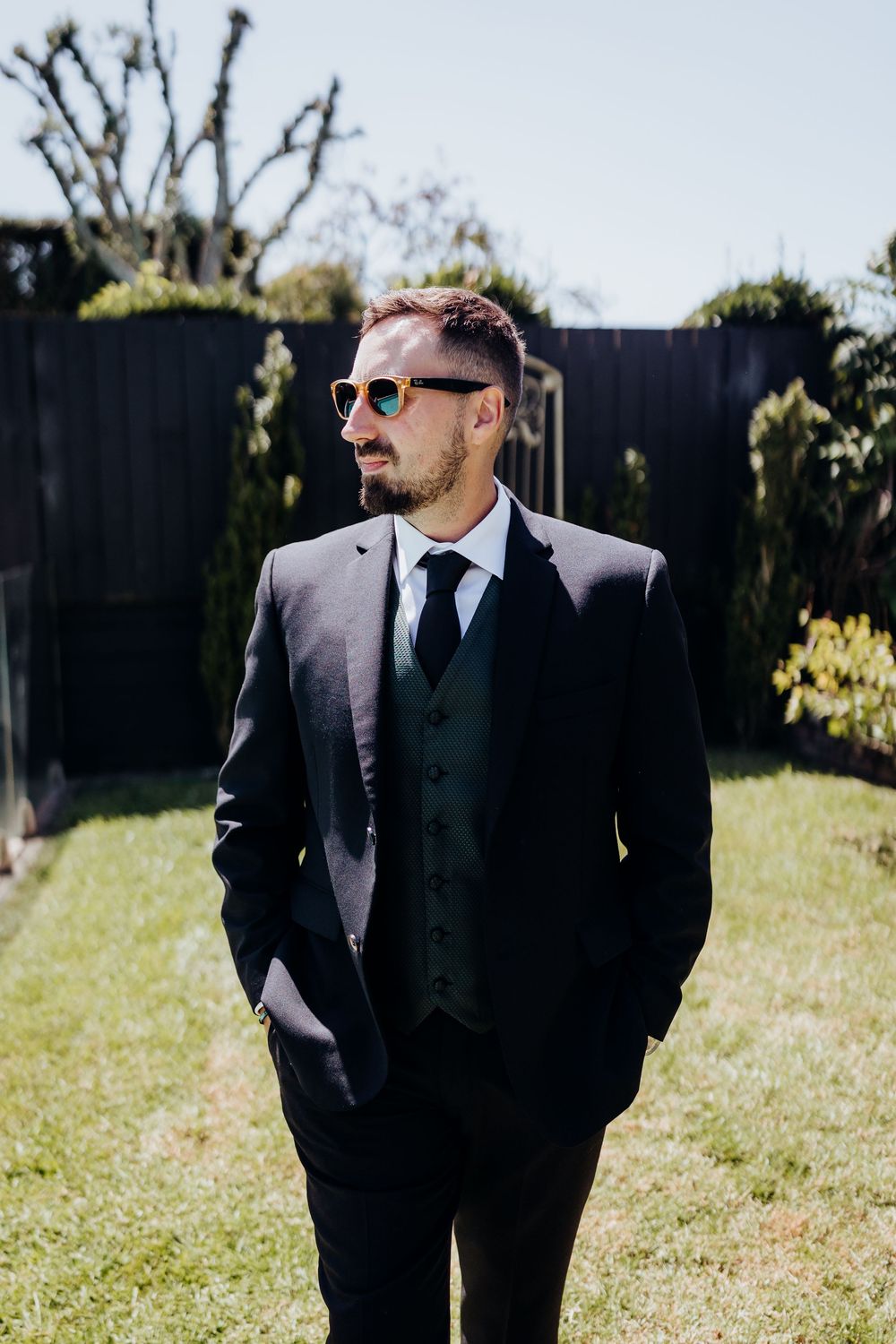 Stylish groom in dark three-piece suit and sunglasses posing outdoors on sunny wedding day.