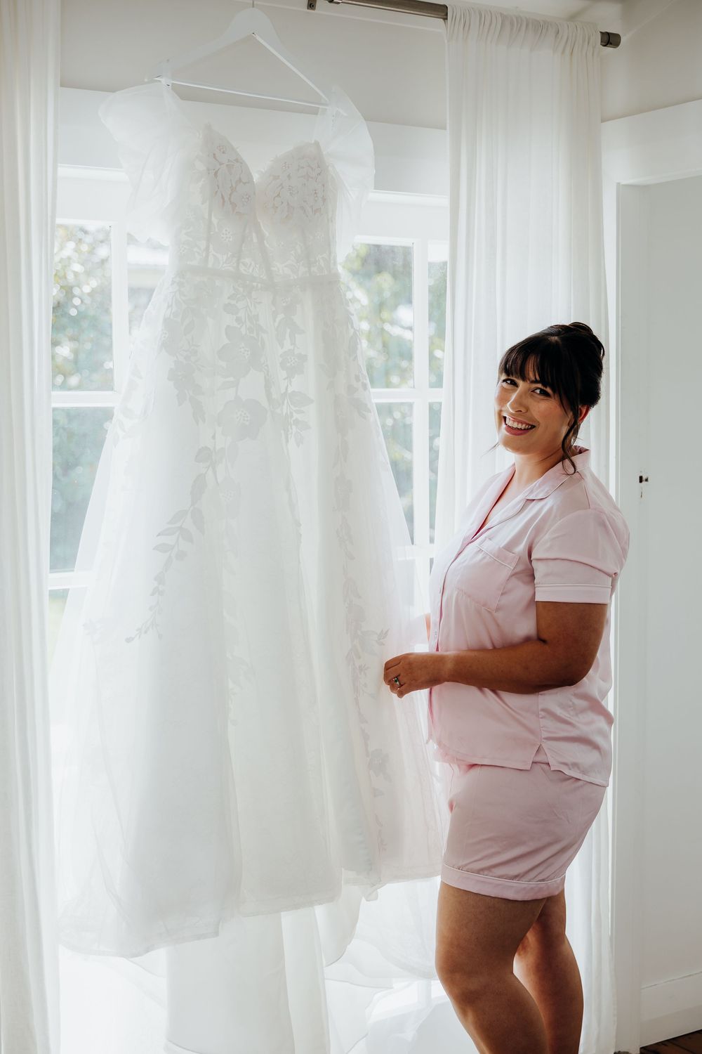 Smiling bride in pink pajamas admiring her lace wedding dress hanging by window.