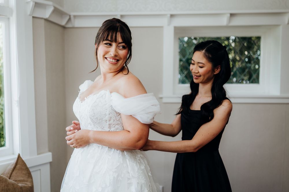 A bride in a white off-shoulder gown smiles as a woman in black helps her dress in a bright room.