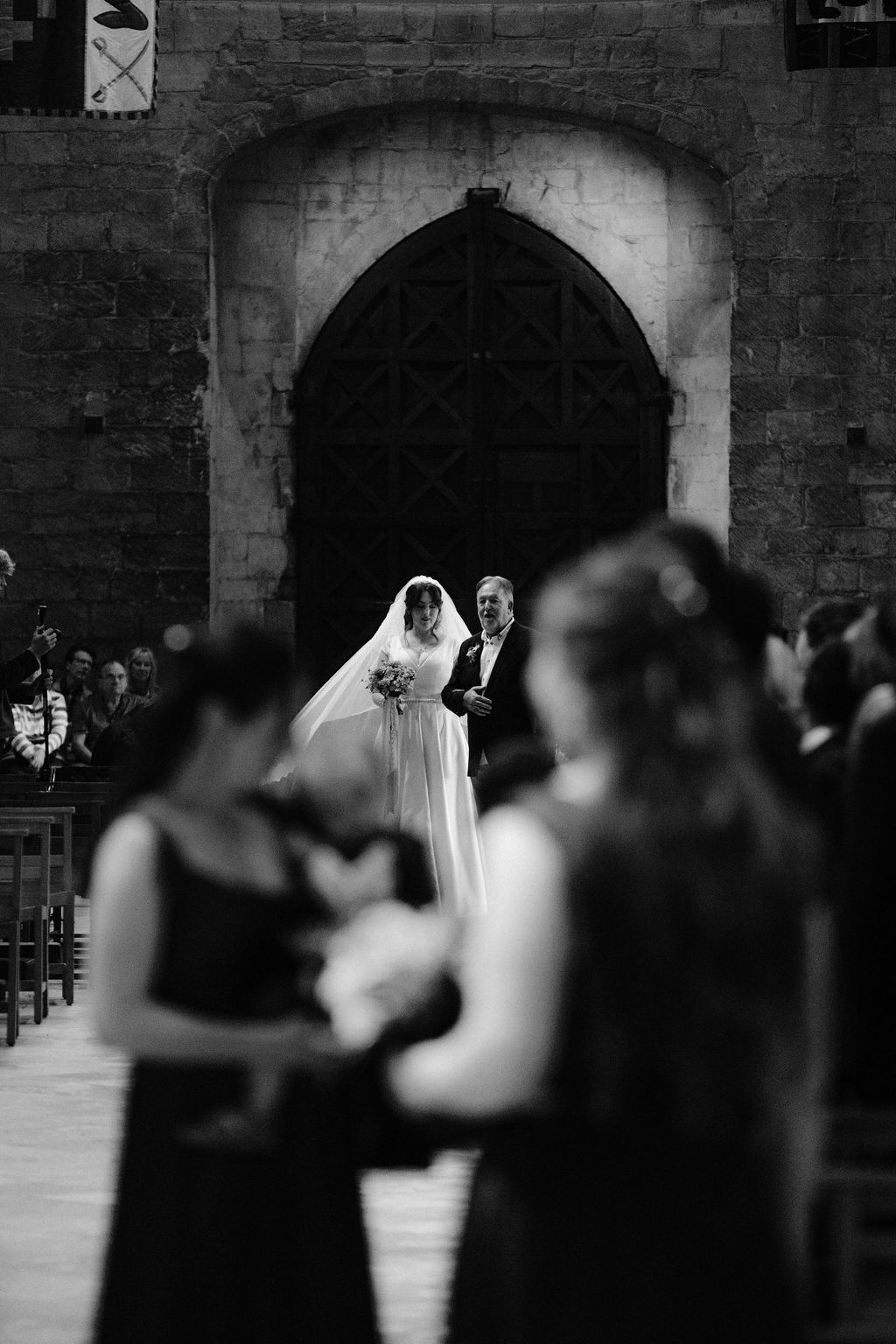 Black and white wedding photo of bride walking down church aisle with escort, bridesmaids in foreground.