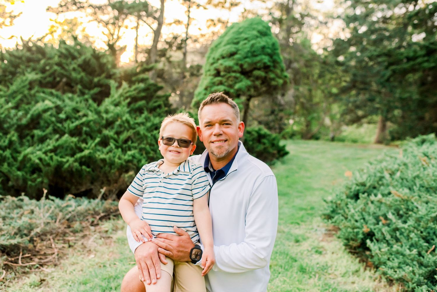 A man holds a young boy wearing sunglasses outdoors in a lush green garden setting during golden hour.