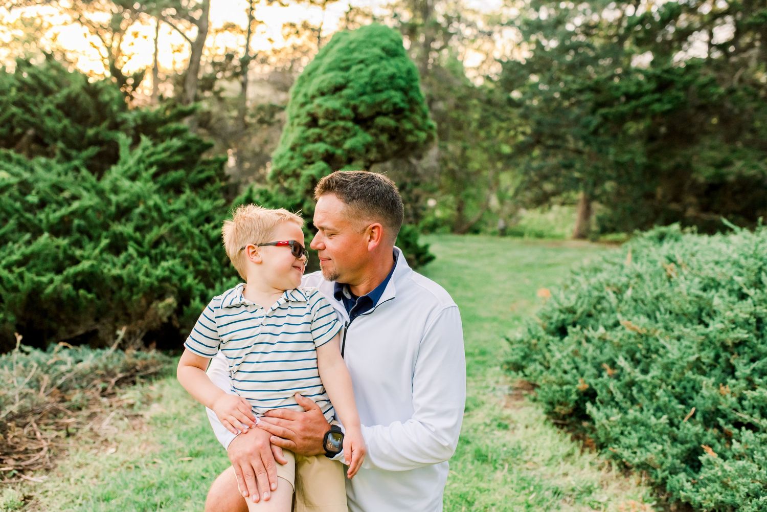 A father holds his young red-haired son in a lush green garden, both smiling at each other warmly.