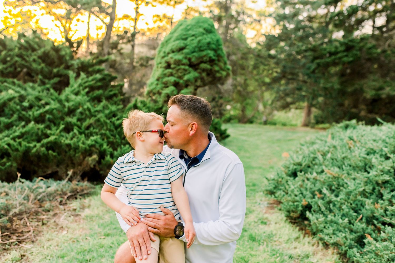 Father kissing young boy in striped shirt outdoors among lush green trees during golden hour.