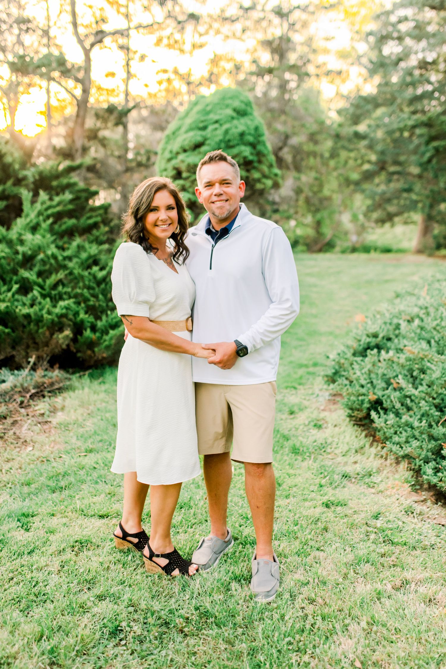 Couple in matching white outfits smiling together on green lawn surrounded by lush garden trees at golden hour. West Lafayette, Indiana.