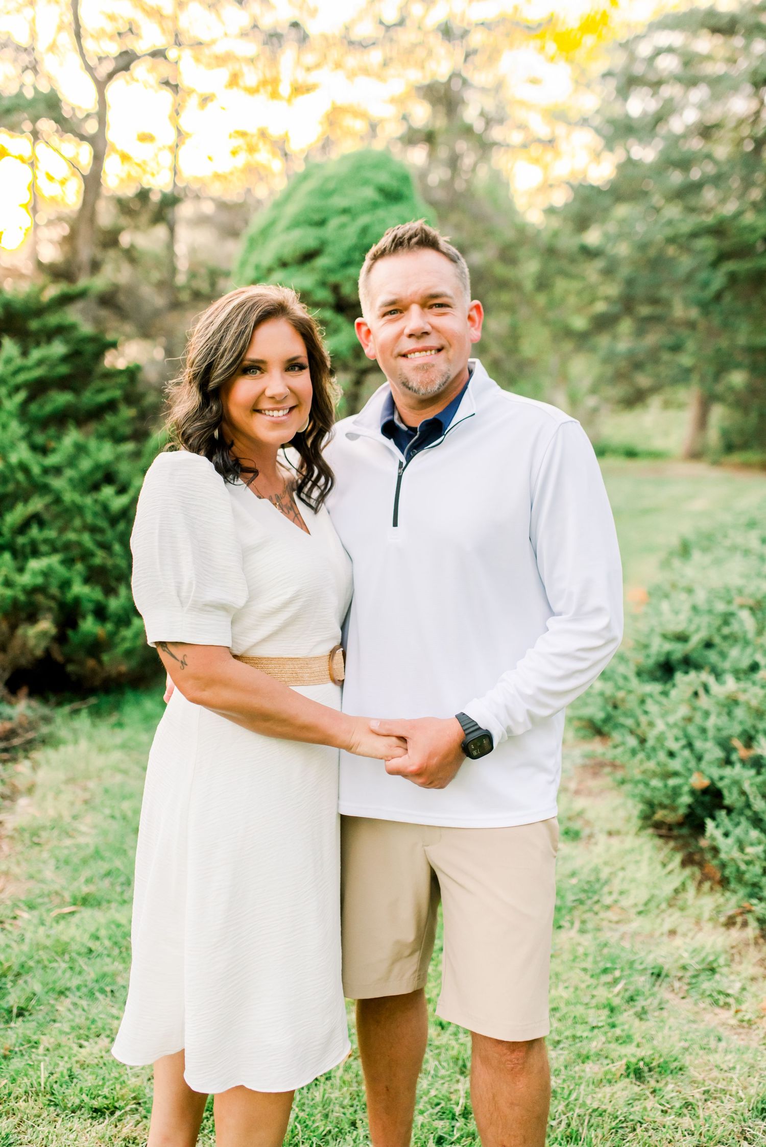 Smiling couple posing together outdoors in a lush garden, woman in white dress and man in khaki shorts.