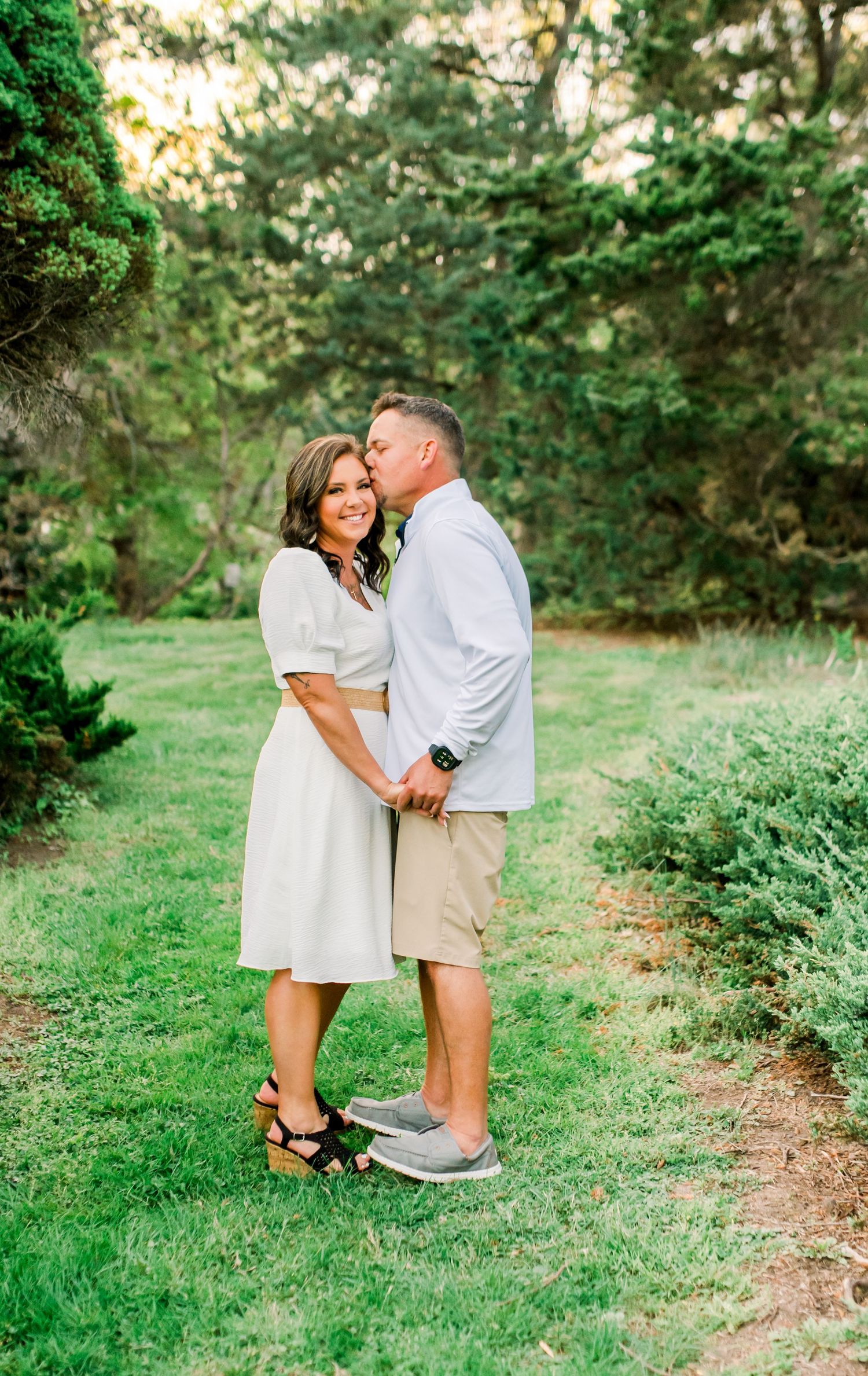 Couple sharing a romantic kiss in a lush green garden, woman in white dress and man in light blue shirt.