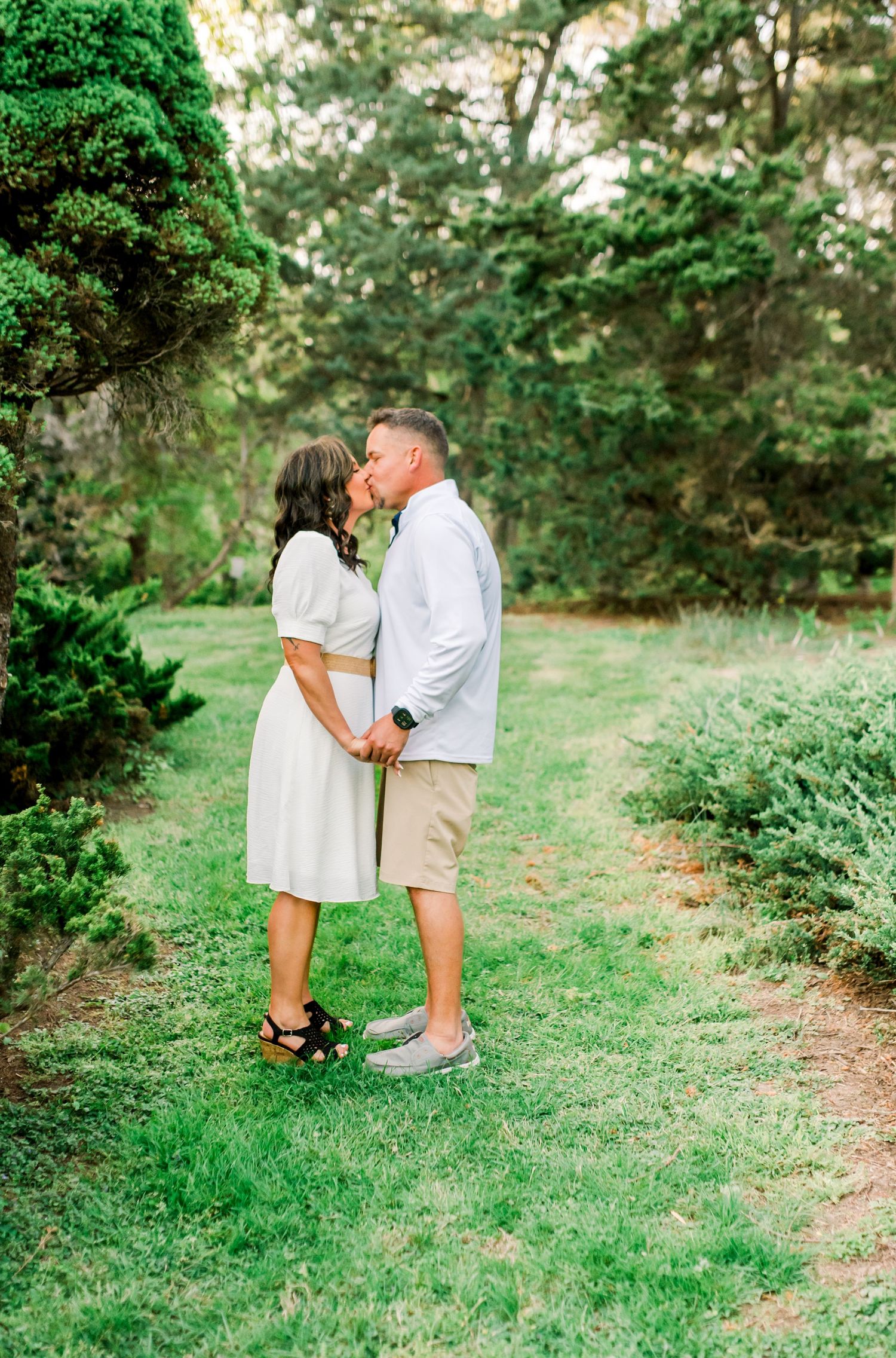 Couple sharing a romantic kiss while holding hands on a green lawn surrounded by evergreen trees.