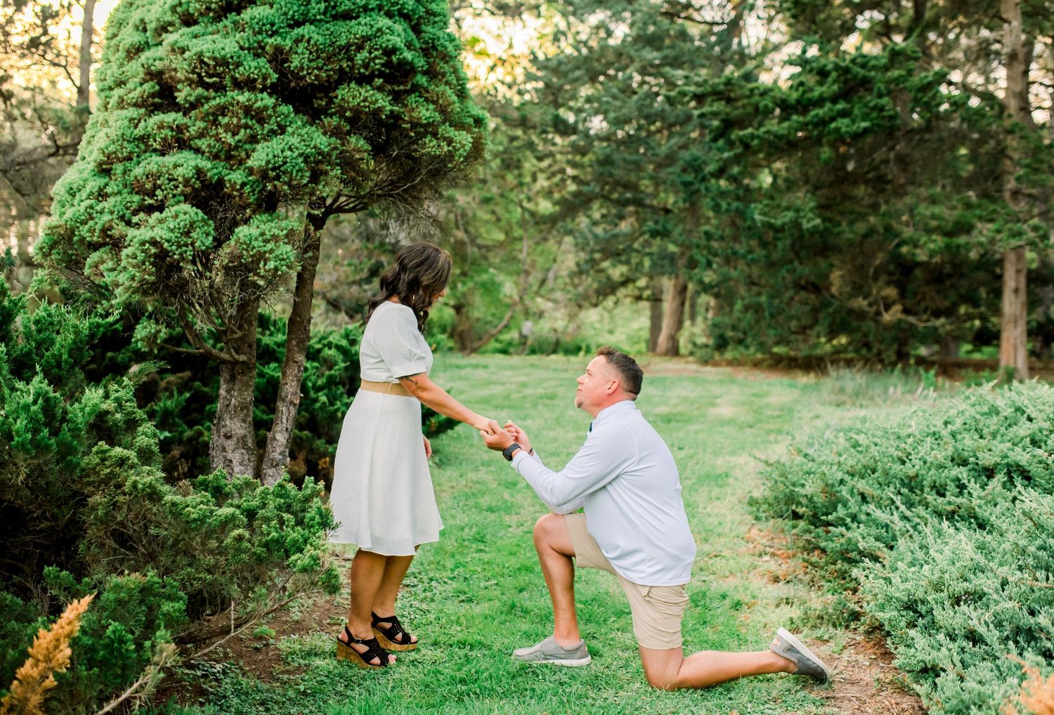 Man proposing on one knee to woman in white dress outdoors in lush green garden setting. Purdue University.