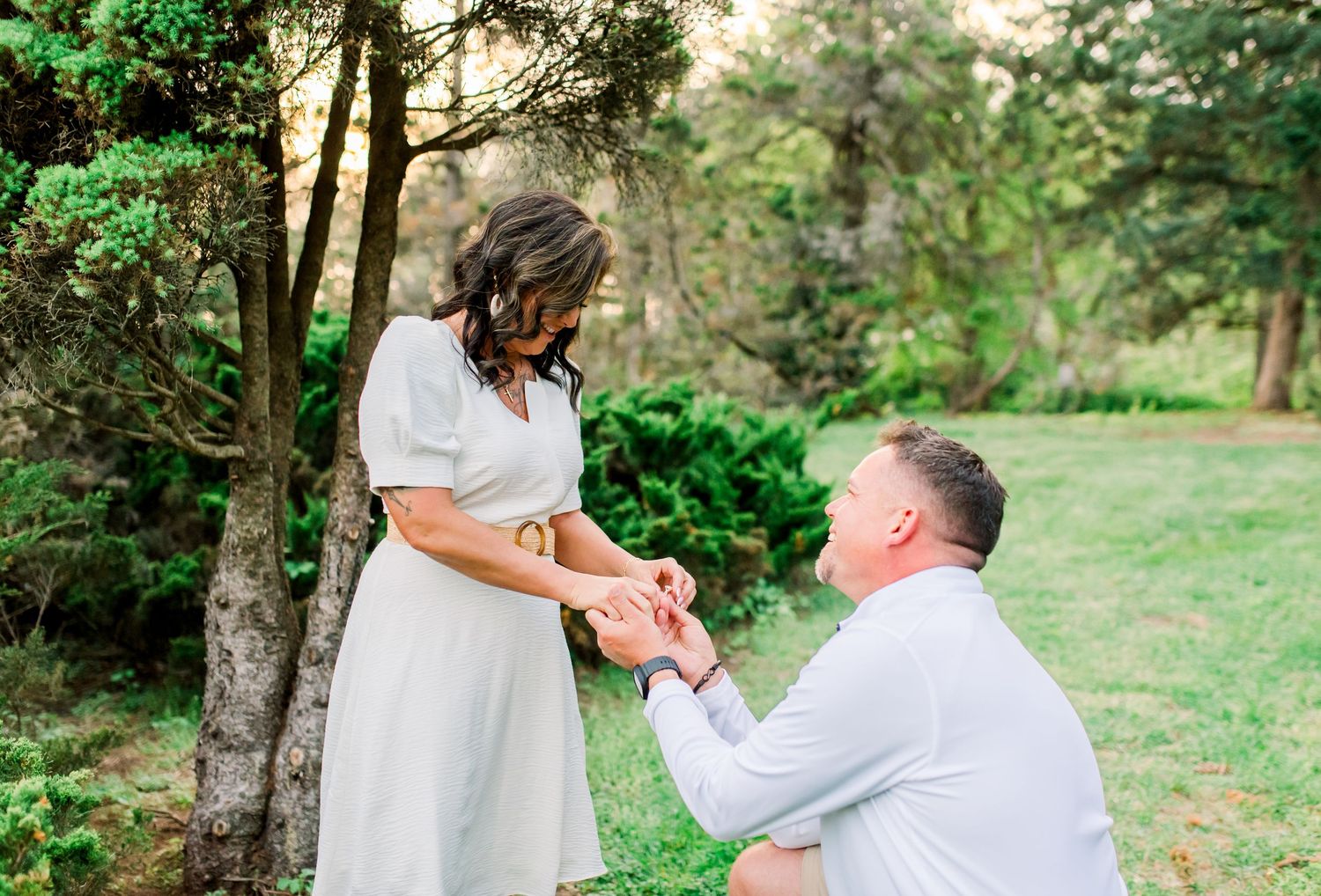 Man kneeling on one knee proposing to woman in white dress outdoors in lush green garden setting.