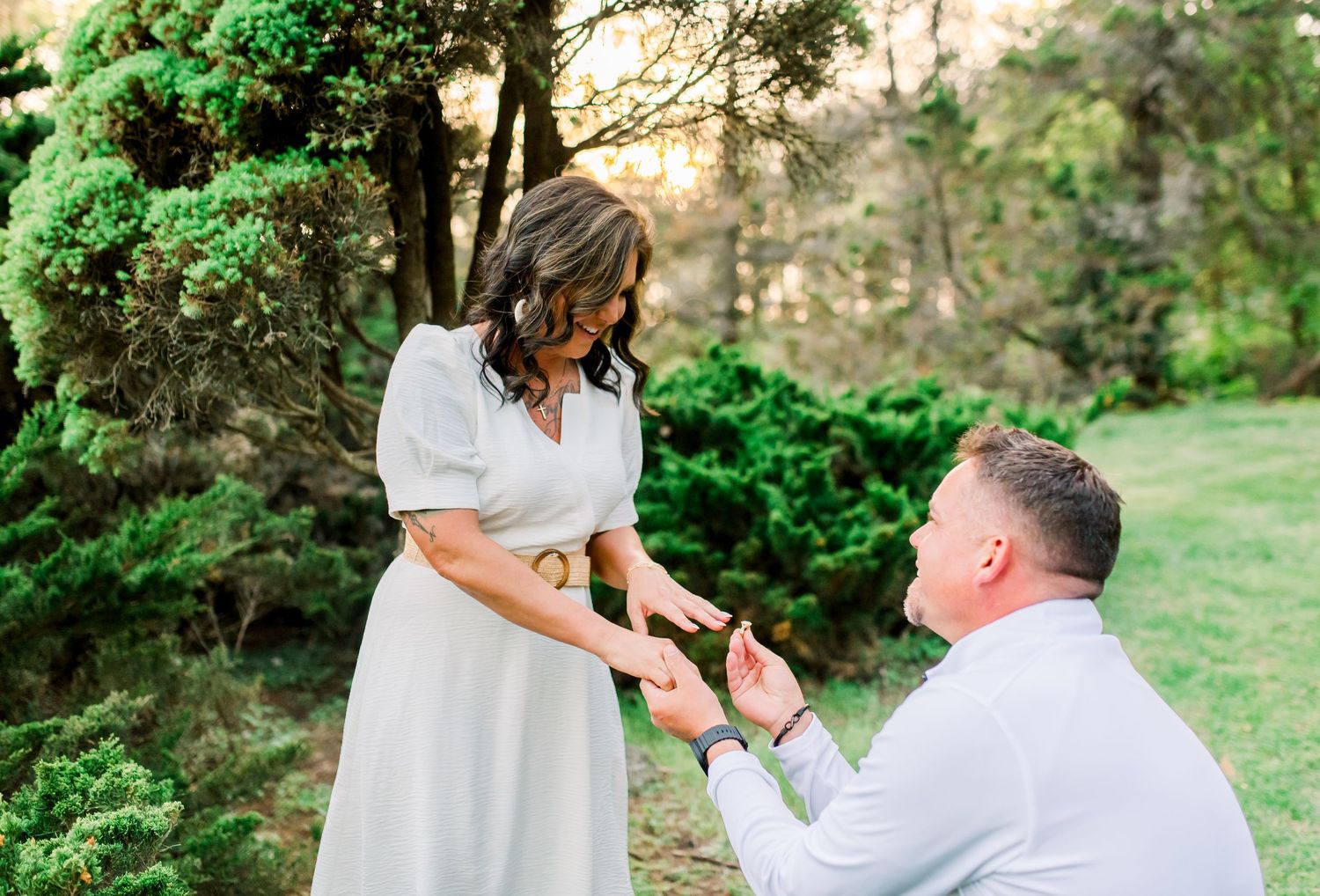 Man kneeling on one knee proposing to woman in white dress outdoors in lush green garden at golden hour.