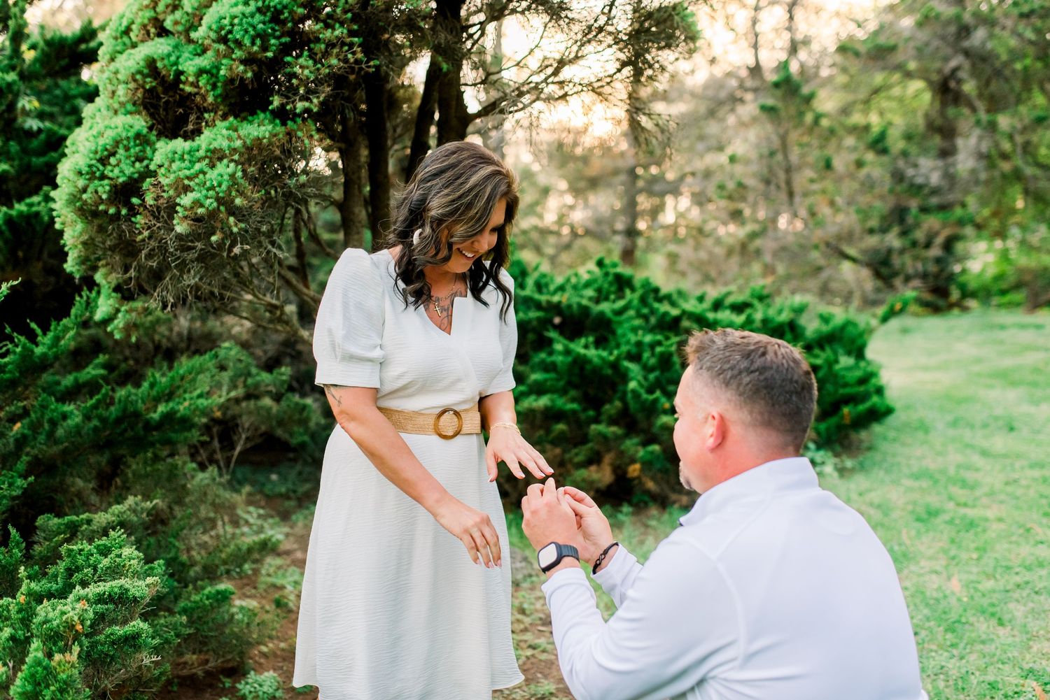 Man kneeling on one knee proposing to a smiling woman in a white dress outdoors at golden hour.