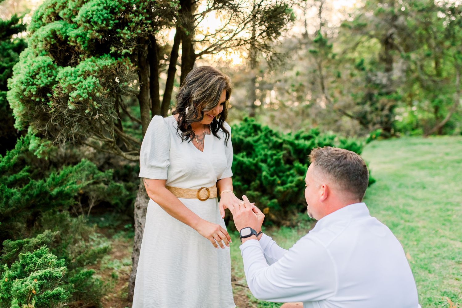 Romantic outdoor marriage proposal with man on one knee placing ring on womans finger in garden setting.