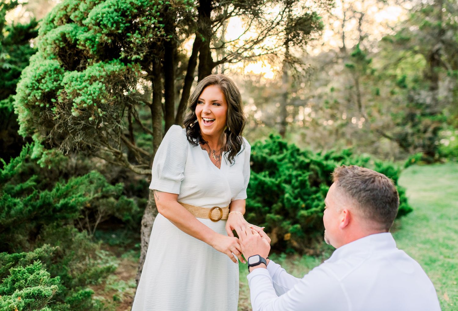 Woman laughing joyfully as man kneels holding her hands during an outdoor marriage proposal at sunset.